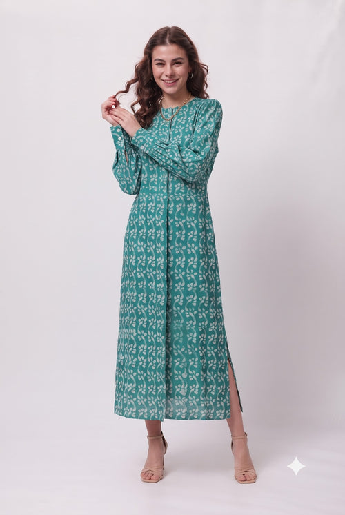 A woman modeling a long-sleeved, floor-length teal green dress with a white geometric pattern and side slits, standing against a light gray studio background.