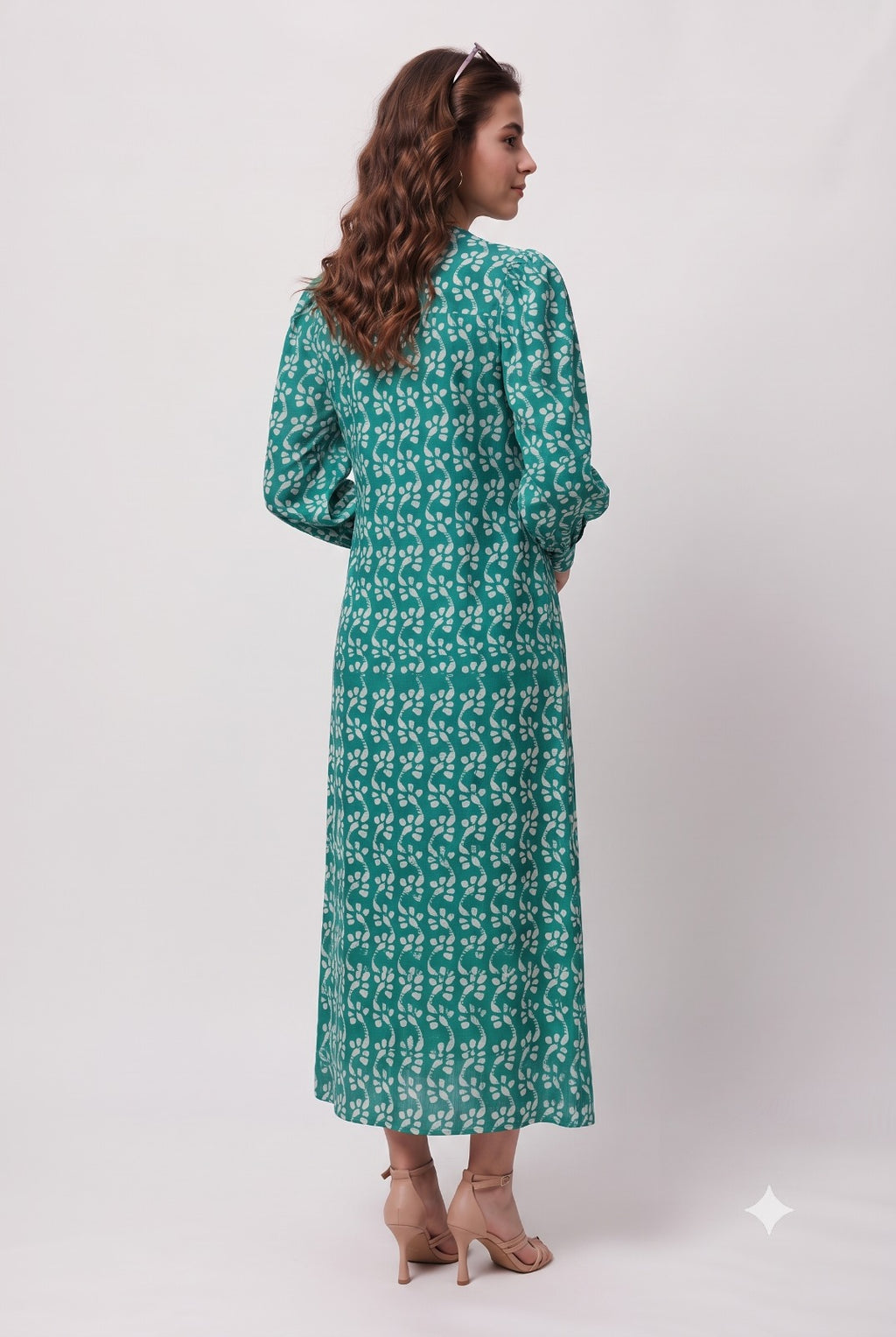 A woman wearing a long-sleeved green maxi dress with a white floral pattern, shown from the back against a plain light gray studio background.