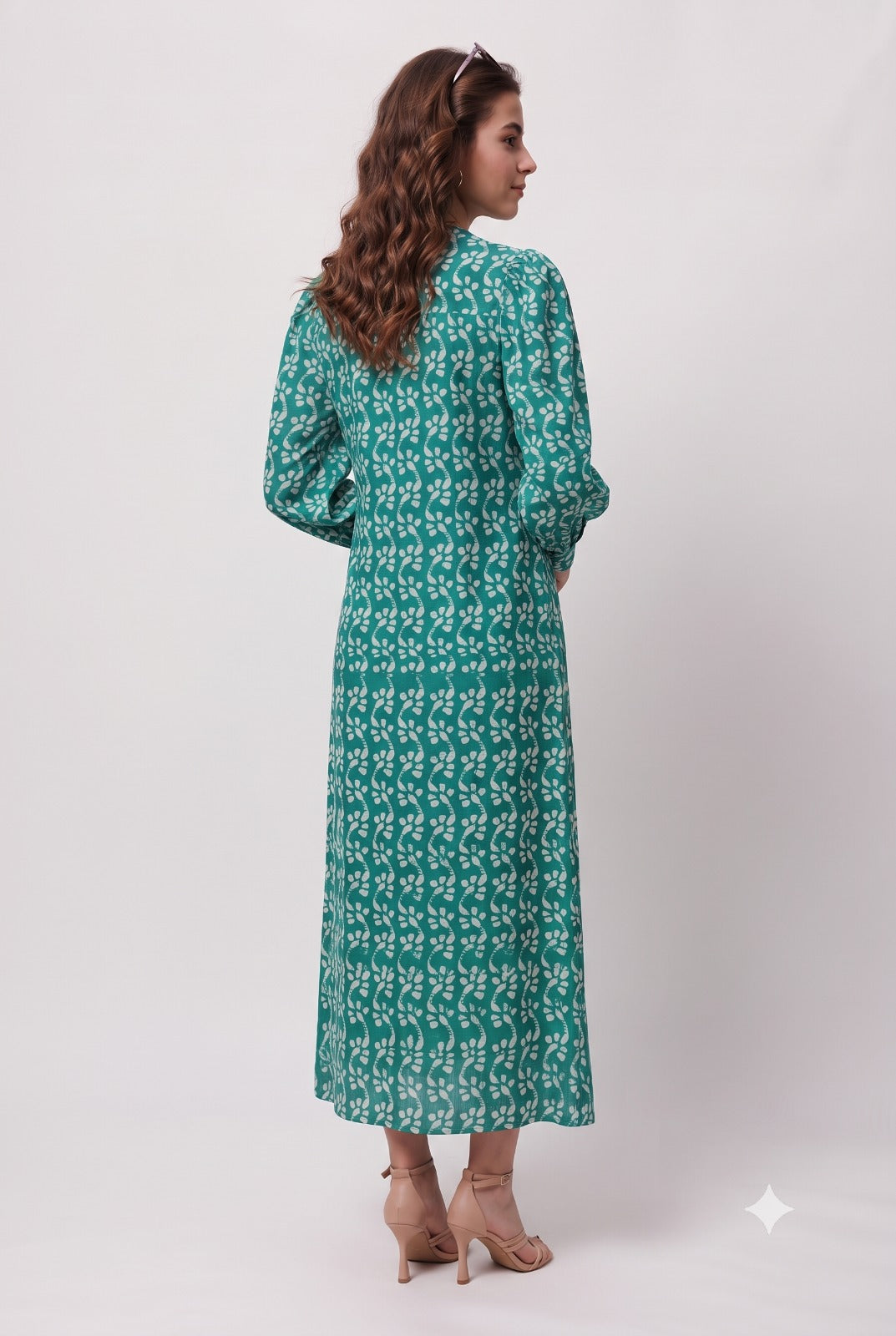 A woman wearing a long-sleeved green maxi dress with a white floral pattern, shown from the back against a plain light gray studio background.