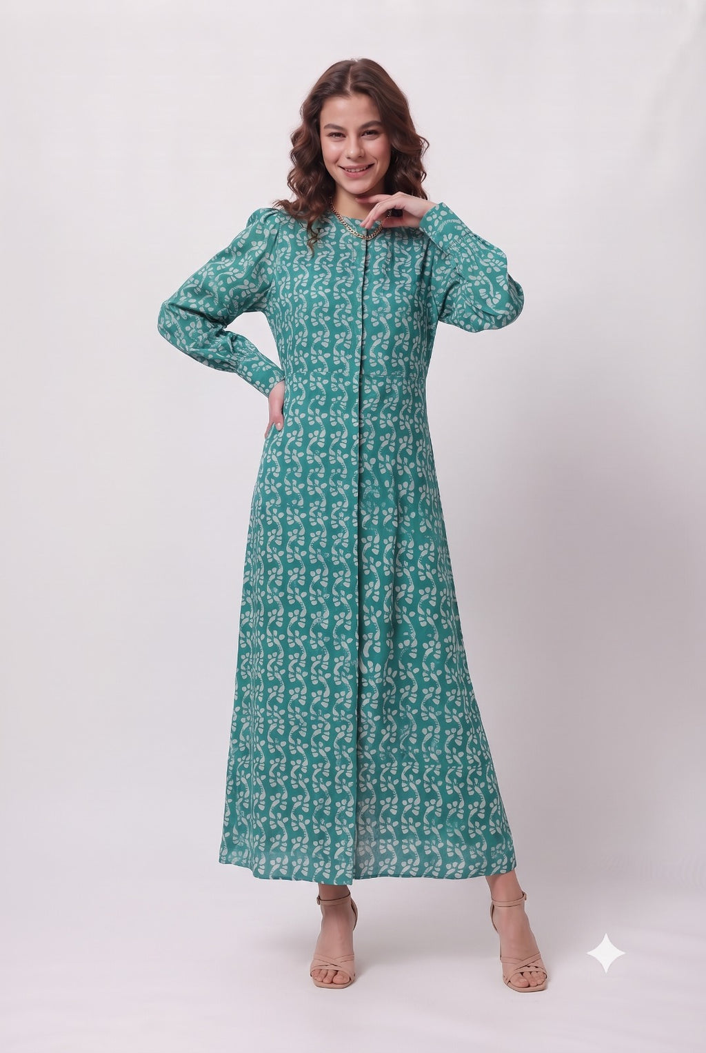 A woman modeling a long-sleeved, floor-length teal green dress with a white geometric pattern, standing against a light gray studio background.