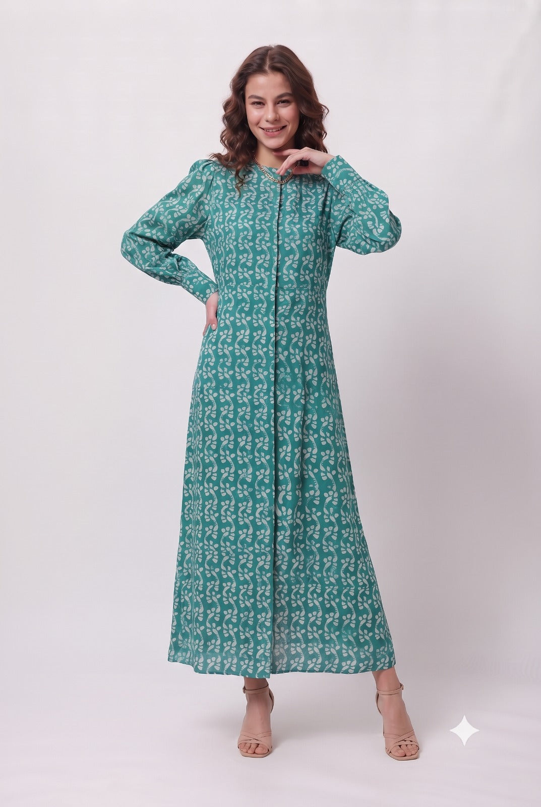 A woman modeling a long-sleeved, floor-length teal green dress with a white geometric pattern, standing against a light gray studio background.