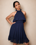 A young girl modeling a navy-blue halter-neck dress with ruffle details, standing against a plain beige studio background.