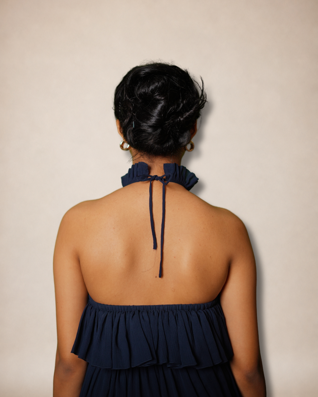 Back view of a woman wearing a navy blue halter top with a ruffled neck and tie detail, set against a plain beige background.