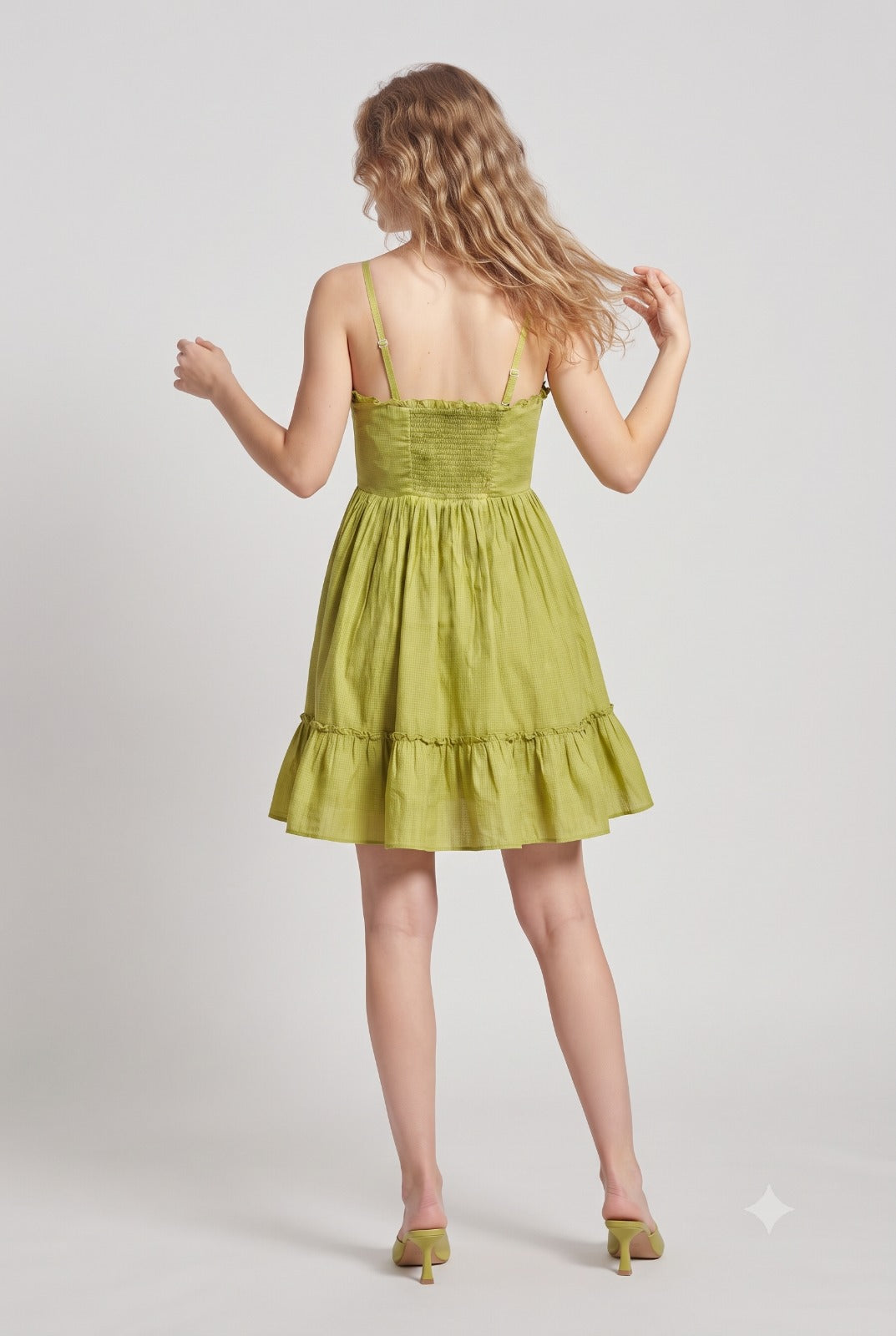 A woman wearing a light olive green tiered mini dress with thin spaghetti straps, shown from the back against a light gray studio background.