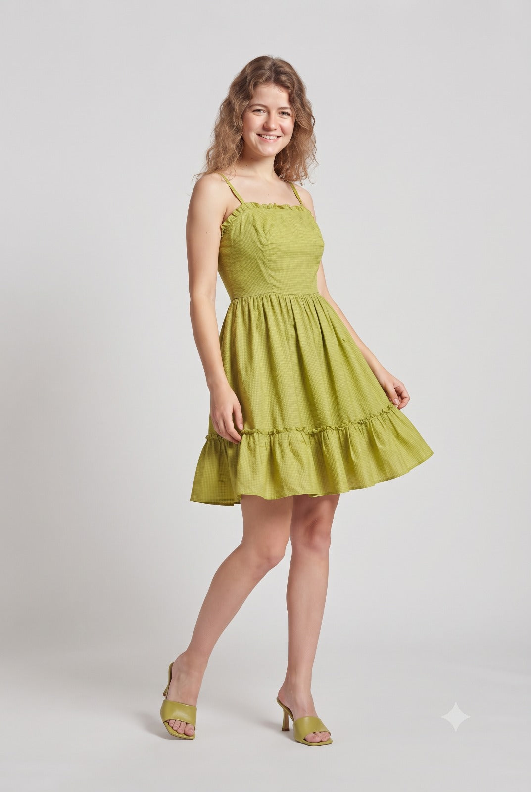 A woman modeling a short olive green tiered dress with thin straps and a flared skirt, paired with matching green heeled sandals, against a light gray studio background.