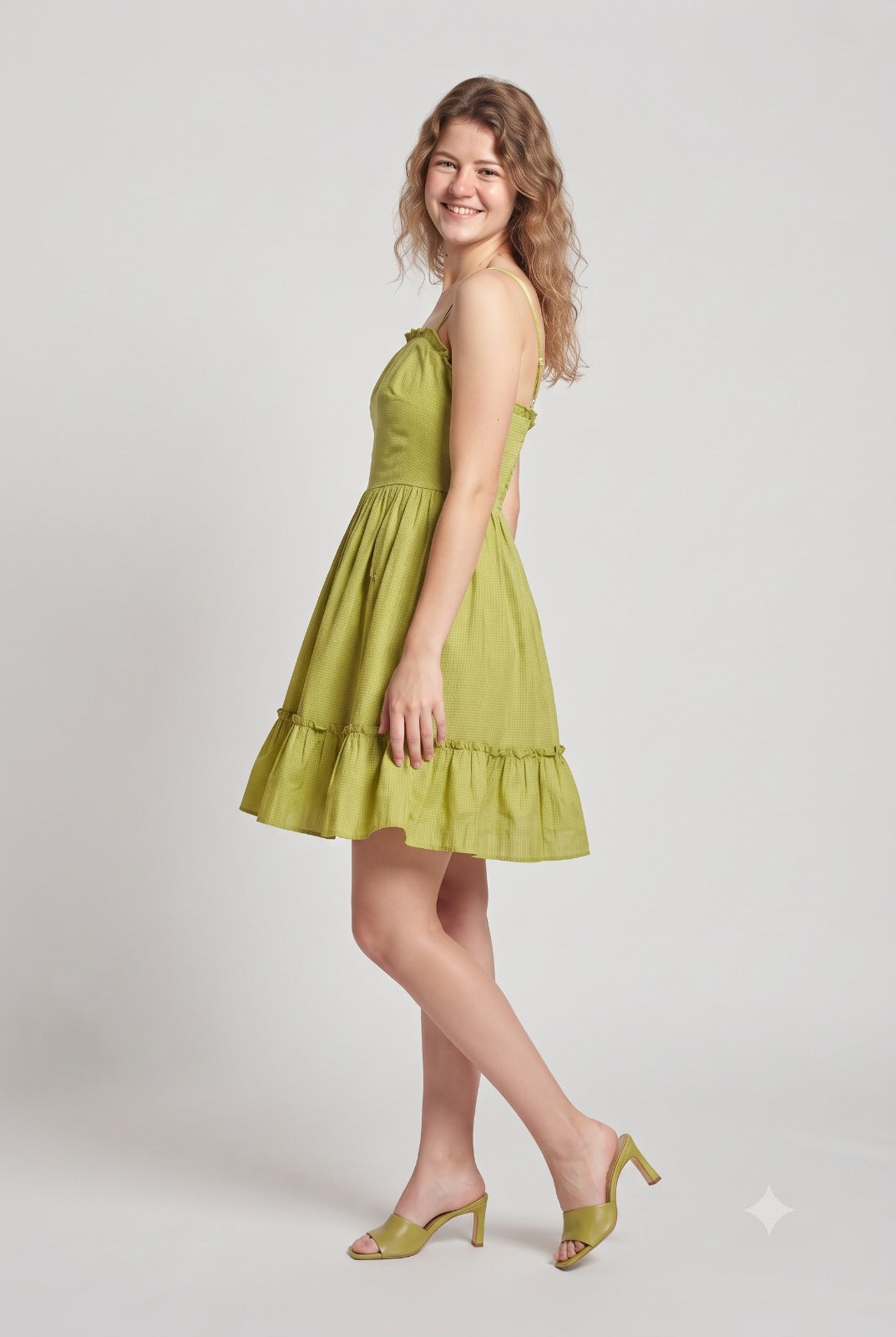 A woman modeling a sleeveless olive green tiered mini dress with ruffled trim, paired with matching olive green heeled sandals, against a light gray studio background.