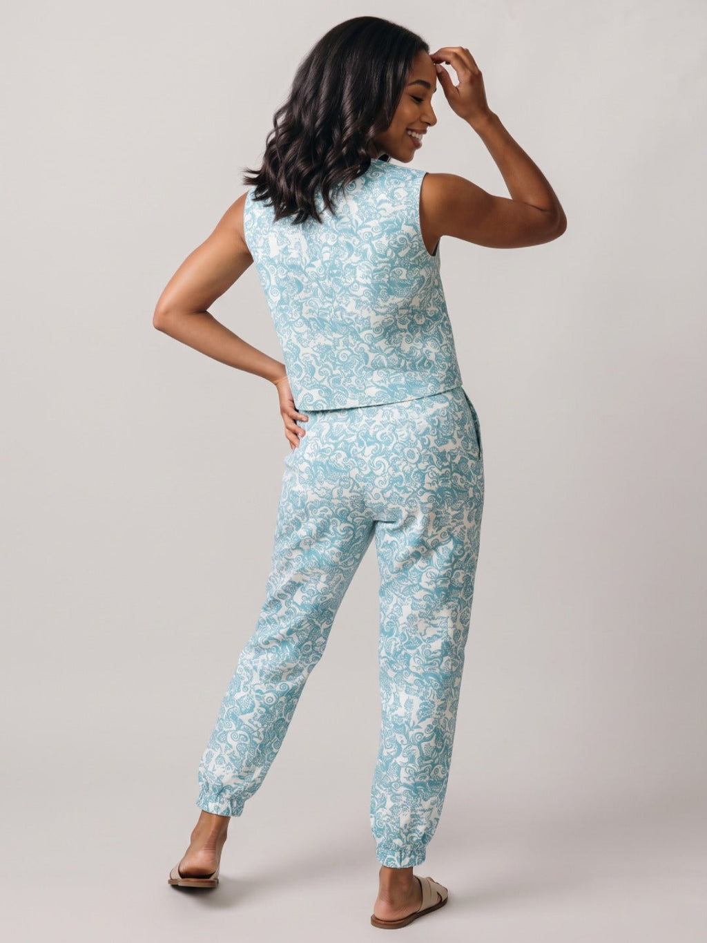 A woman modeling a sleeveless light blue and white patterned jumpsuit, shown from a back-three-quarter angle against a neutral light gray studio background.