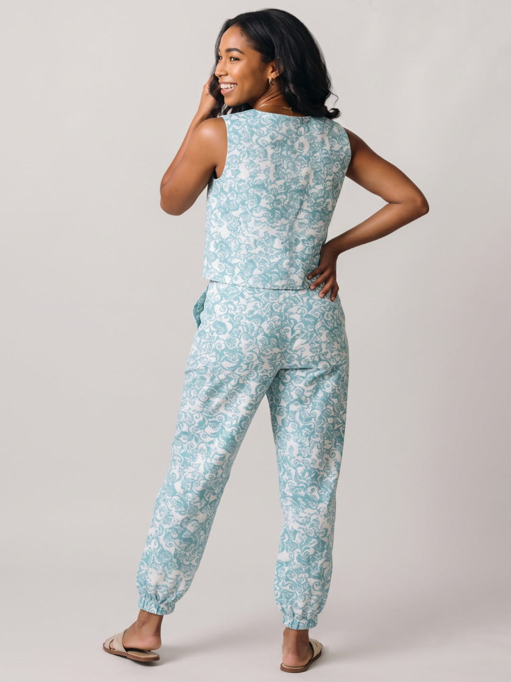 A woman modeling a sleeveless light blue and white floral print jumpsuit, shown from a back-three-quarter angle against a plain light gray studio background.
