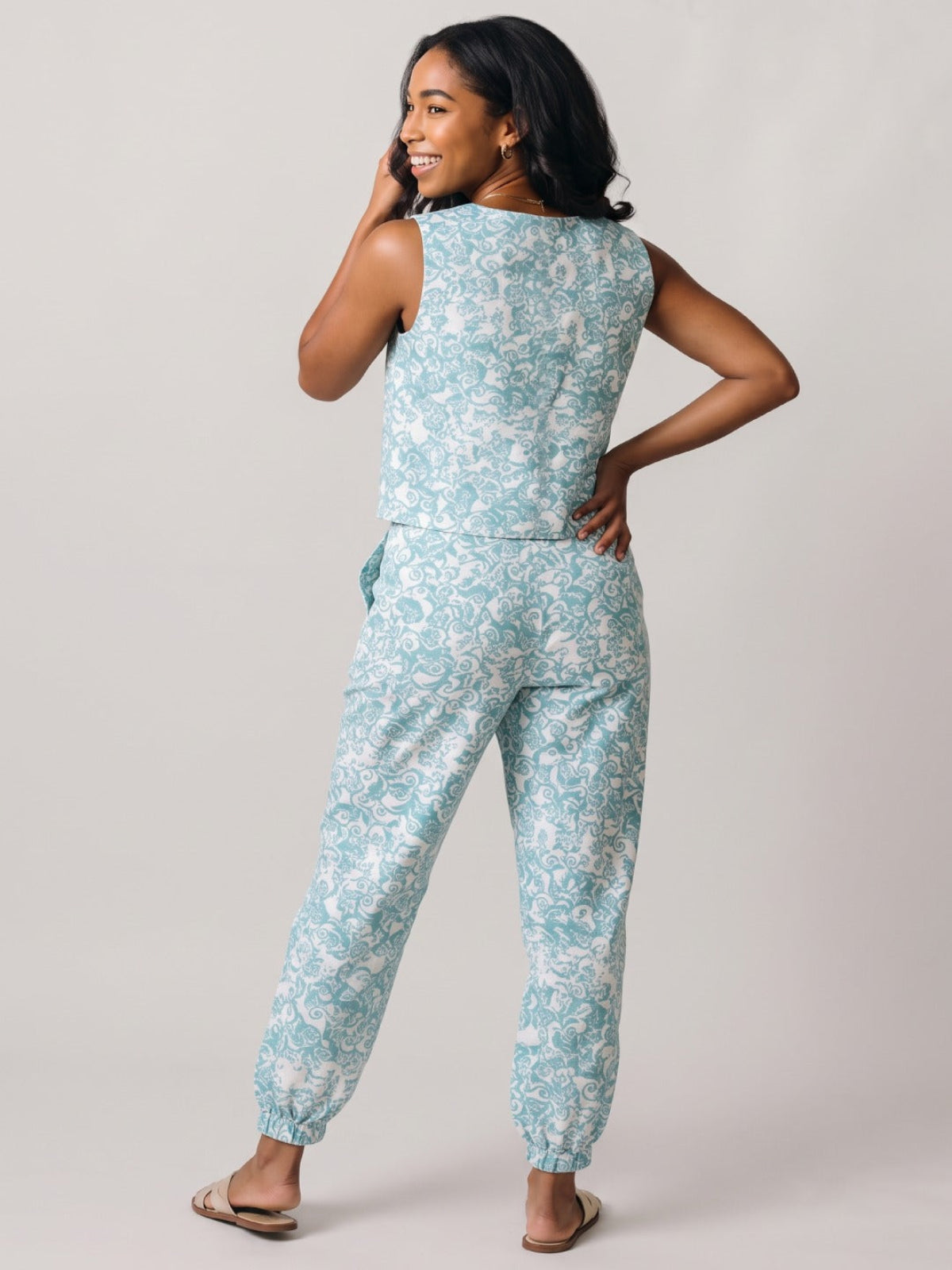 A woman modeling a sleeveless light blue and white floral print jumpsuit, shown from a back-three-quarter angle against a plain light gray studio background.