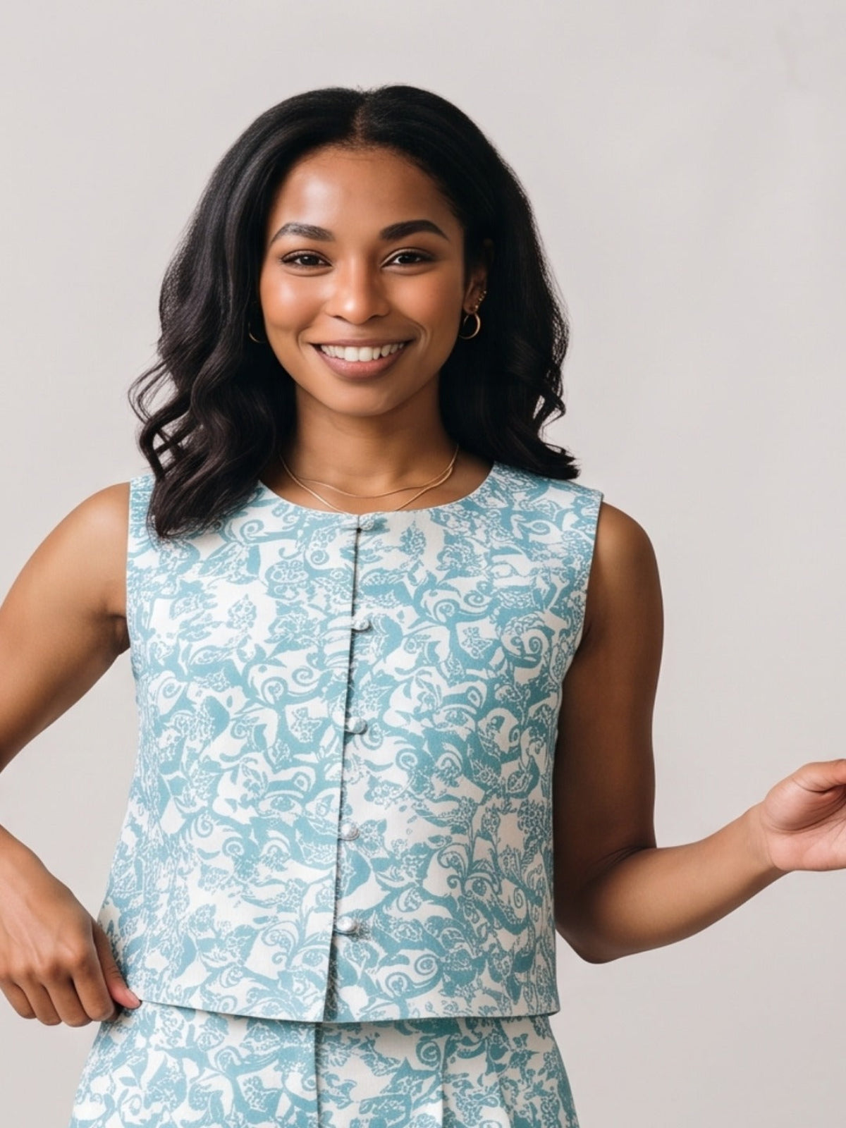 A smiling woman with dark wavy hair wearing a sleeveless light blue and white patterned vest with a button front, posed against a plain light gray studio background.