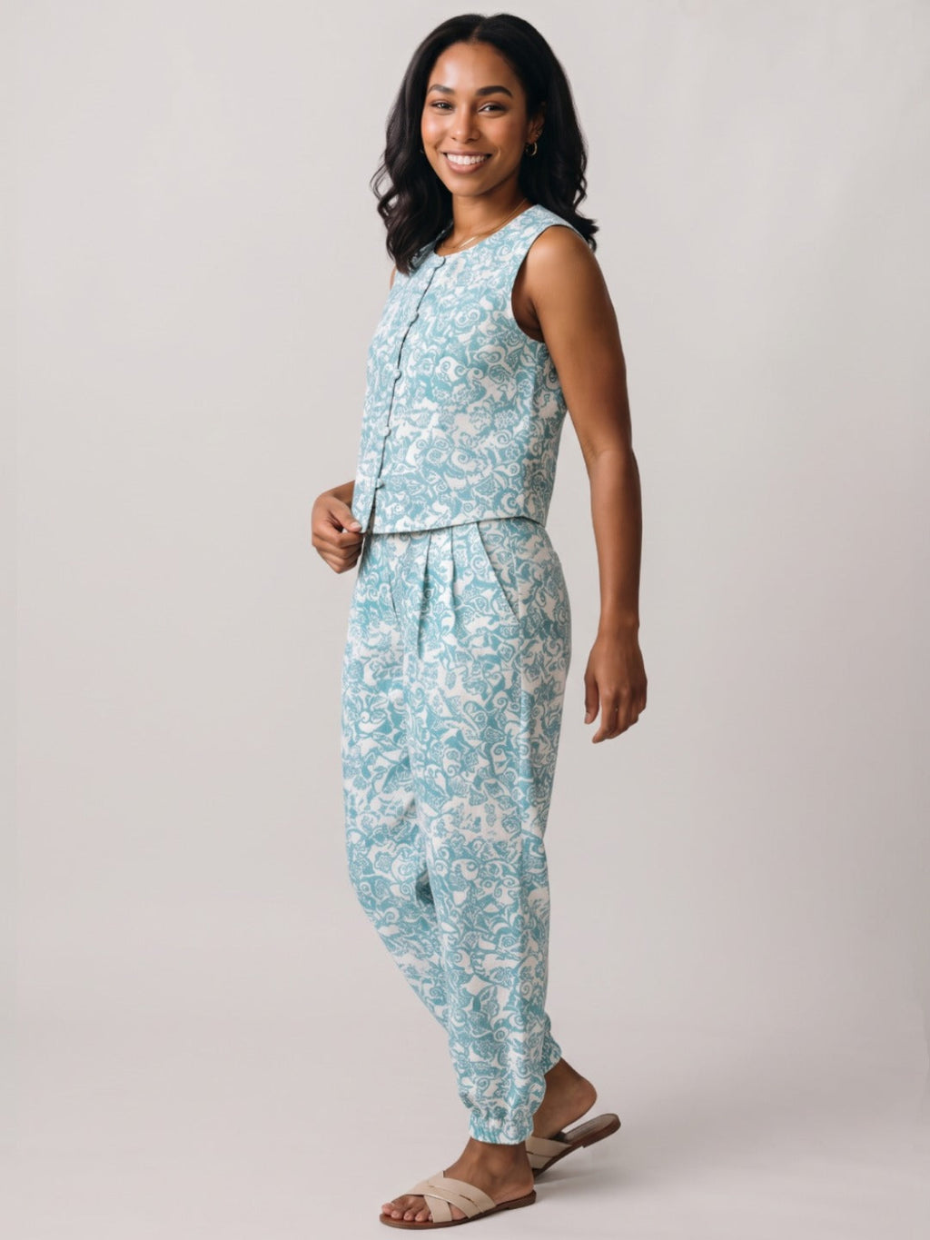 A woman modeling a sleeveless light blue and white patterned jumpsuit against a plain light gray studio background.
