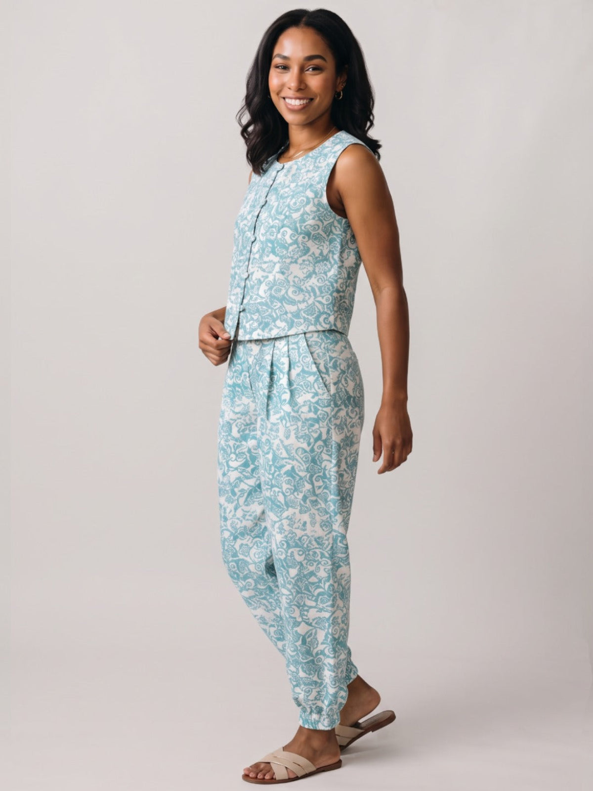 A woman modeling a sleeveless light blue and white patterned jumpsuit against a plain light gray studio background.